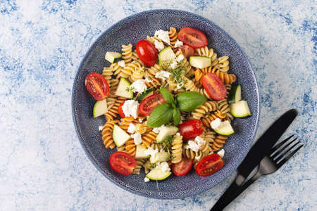 Italian pasta fusilli tricolore with cherry tomatoes, zucchini and feta cheese in a blue shallow bow with black silverware on a blue surfaceの写真素材