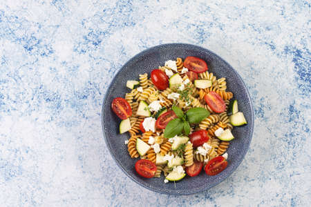 Italian pasta fusilli tricolore with cherry tomatoes, zucchini and feta cheese in a blue shallow bowl on a blue surfaceの写真素材