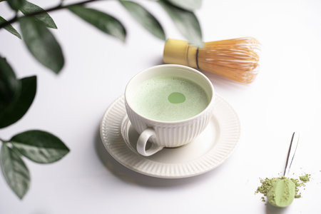 A white porcelain cup with japanese matcha tea drink on a white saucer plate on a white surface, a tea spoon with matcha powder, bamboos whisker and a green plant zamioculcasの写真素材
