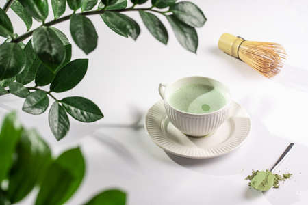 A white porcelain cup with japanese matcha tea drink on a white saucer plate on a white surface, a tea spoon with matcha powder, bamboos whisker and a green plant zamioculcasの写真素材