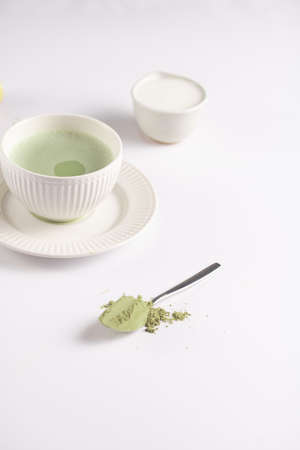 A white porcelain cup with japanese matcha tea drink on a white saucer plate on a white surface, a tea spoon with matcha powder, bamboos whiskerの写真素材