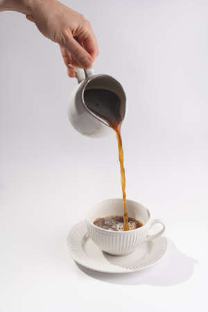 Female hand pouring coffee into a white porcelain cup with coffee drink cappuccino on a white saucer plate on a white surfaceの写真素材
