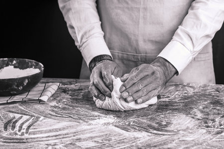 A male chef baker in a white shirt and gray apron kneading dough on a dark kitchen table, black and whiteの写真素材