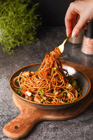 Female hand holding a fork with Italian pasta spaghetti with chicken, green beans, cheese mozzarella and fresh thyme in green bowl on wooden board on gray concrete surfaceの写真素材