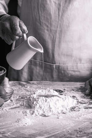 A male chef cook in a white shirt and gray apron pouring water into a heap of flour, black and white