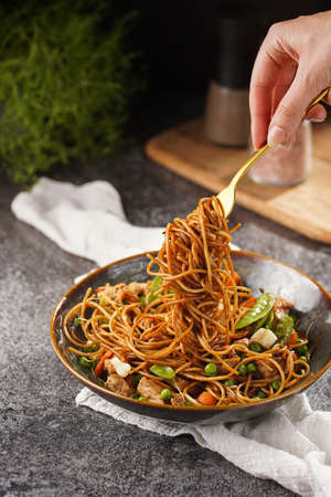 Female hand holding a fork with Italian pasta spaghetti with chicken, green beans, cheese mozzarella and fresh thyme in green bowl on wooden board on gray concrete surfaceの写真素材