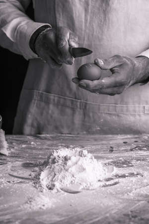 A male chef cook in a white shirt and gray apron breaking an egg into a heap of flour, black and whiteの写真素材