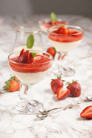 Several glasses with traditional creamy pudding dessert panna cotta with peppermint and strawberry jam surrounded by fresh strawberries on a marble surfaceの写真素材