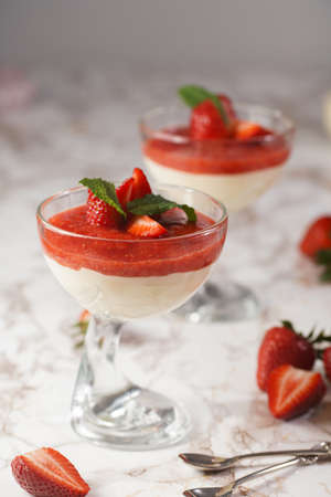 Several glasses with traditional creamy pudding dessert panna cotta with peppermint and strawberry jam surrounded by fresh strawberries on a marble surfaceの写真素材