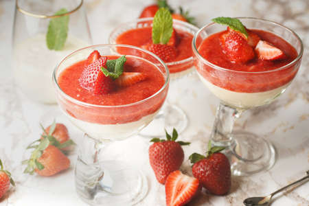 Several glasses with traditional creamy pudding dessert panna cotta with peppermint and strawberry jam surrounded by fresh strawberries on a marble surfaceの写真素材