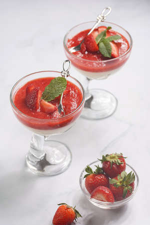 Several glasses with traditional creamy pudding dessert panna cotta with peppermint and strawberry jam surrounded by fresh strawberries on a white surfaceの写真素材