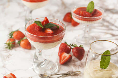 Several glasses with traditional creamy pudding dessert panna cotta with peppermint and strawberry jam surrounded by fresh strawberries on a marble surfaceの写真素材