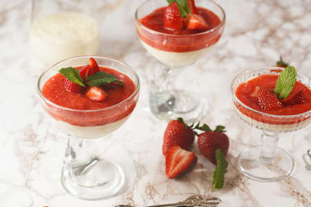 Several glasses with traditional creamy pudding dessert panna cotta with peppermint and strawberry jam surrounded by fresh strawberries on a marble surfaceの写真素材