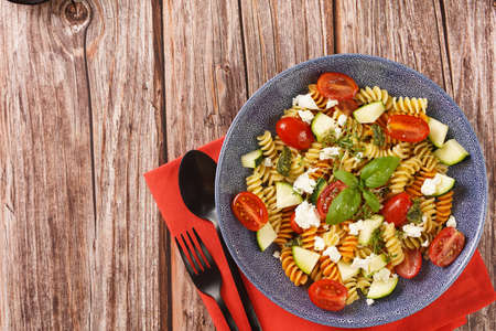 Italian pasta fusilli tricolore with cherry tomatoes, zucchini and feta cheese in a blue shallow bowl on a red napkin with black silverware on a wooden surfaceの写真素材