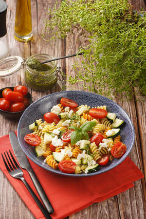Italian pasta fusilli tricolore with cherry tomatoes, zucchini and feta cheese in a blue shallow bowl on a red napkin with black silverware on a wooden surfaceの写真素材