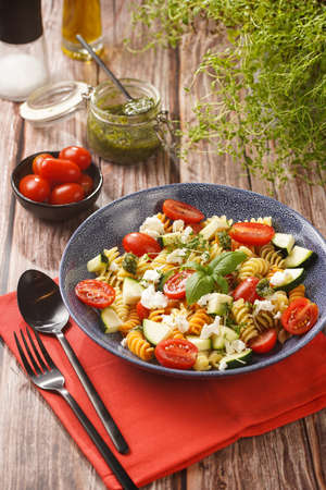 Italian pasta fusilli tricolore with cherry tomatoes, zucchini and feta cheese in a blue shallow bowl on a red napkin with black silverware on a wooden surfaceの写真素材