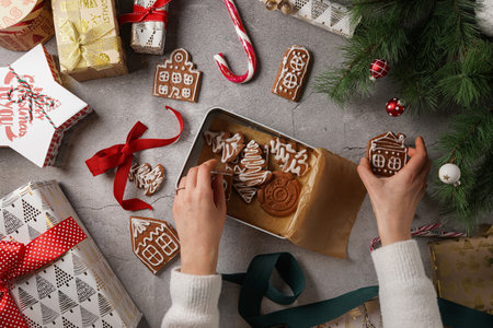 Gingerbread cookies with white sugar frosting shaped like houses on a gray surface surrounded by Christmas decoration and presents and female hands holding a box with cookiesの写真素材