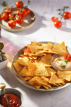 A gray plate with cheese tortilla chips and a chilli cheese dip on a gray surface, cherry tomatoes, ketchup in hard morning lightの写真素材