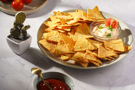 A gray plate with cheese tortilla chips and a chilli cheese dip on a gray surface, cherry tomatoes, ketchup in hard morning lightの写真素材