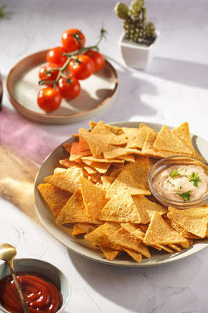 A gray plate with cheese tortilla chips and a chilli cheese dip on a gray surface, cherry tomatoes, ketchup in hard morning lightの写真素材