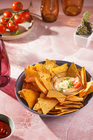 A blue bowl with cheese tortilla chips and a chilli cheese dip on a pink marble surface, cherry tomatoes, ketchup in hard morning lightの写真素材