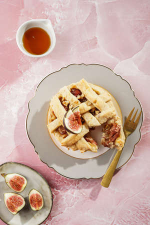 A sweet fig tart pastry on a sophisticated plate, two golden dessert spoons, honey, cut figs on a pink marble surfaceの写真素材