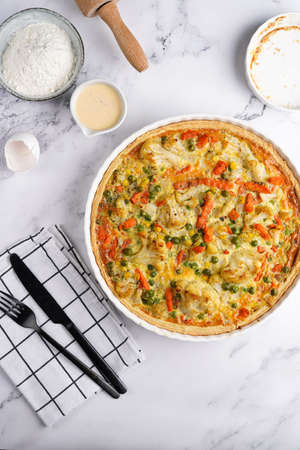 Baked french vegetable pie quiche filled with carrots, peas and cheese on a marble surface, cracked egg shell, flour, cream, top viewの写真素材