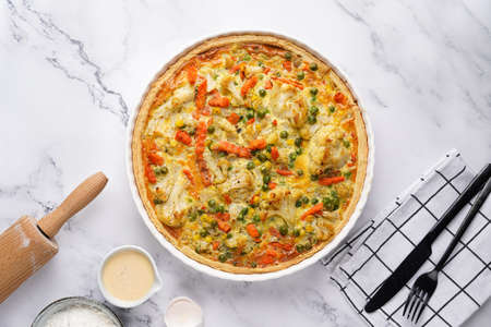 Baked french vegetable pie quiche filled with carrots, peas and cheese on a marble surface, cracked egg shell, flour, cream, top viewの写真素材