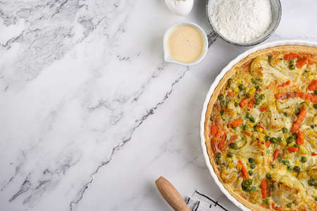 Baked french vegetable pie quiche filled with carrots, peas and cheese on a marble surface, cracked egg shell, flour, cream, top viewの写真素材