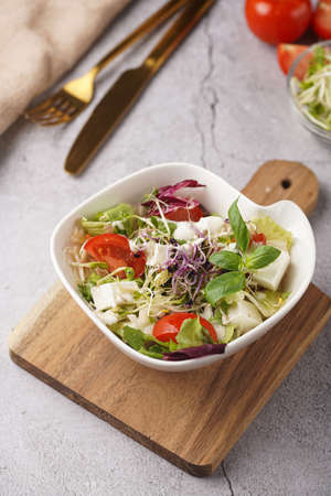 A small white salad bowl with microgreens, cherry tomatoes and fresh basil leaves on a wooden board on a gray concrete surfaceの写真素材