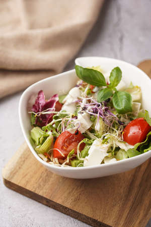 A small white salad bowl with microgreens, cherry tomatoes and fresh basil leaves on a wooden board on a gray concrete surfaceの写真素材
