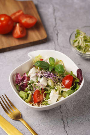 A small white salad bowl with microgreens, cherry tomatoes and fresh basil leaves on a wooden board on a gray concrete surfaceの写真素材