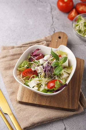 A small white salad bowl with microgreens, cherry tomatoes and fresh basil leaves on a wooden board on a gray concrete surfaceの写真素材