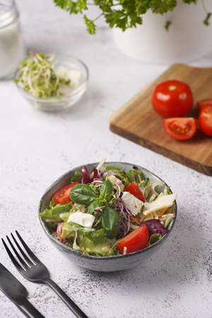 A small white salad bowl with microgreens, cherry tomatoes and fresh basil leaves on a wooden board on a gray concrete surfaceの写真素材