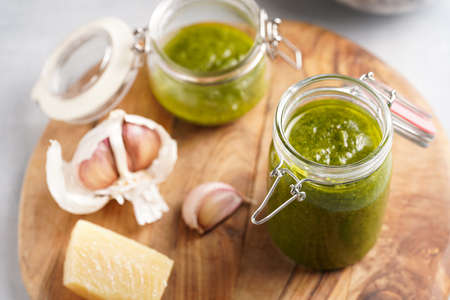 Several small glass mason jars with green italian basil sauce pesto on a round wooden tray with hard cheese pecorino and garlic clovesの写真素材