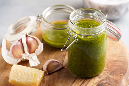 Several small glass mason jars with green italian basil sauce pesto on a round wooden tray with hard cheese pecorino and garlic clovesの写真素材
