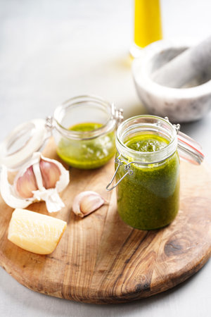 Several small glass mason jars with green italian basil sauce pesto on a round wooden tray with hard cheese pecorino and garlic clovesの写真素材