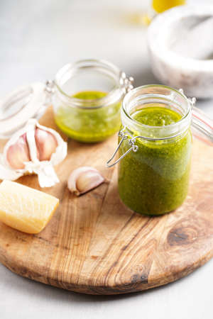 Several small glass mason jars with green italian basil sauce pesto on a round wooden tray with hard cheese pecorino and garlic clovesの写真素材