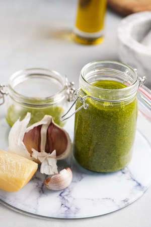 Several small glass mason jars with green italian basil sauce pesto on a round marble tray with hard cheese pecorino and garlic clovesの写真素材