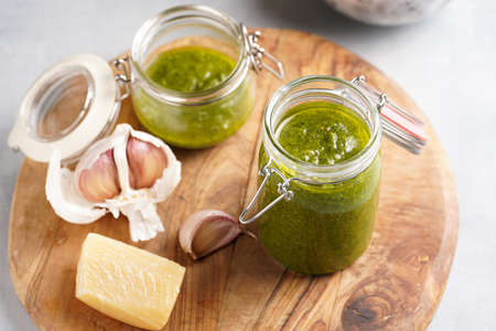 Several small glass mason jars with green italian basil sauce pesto on a round wooden tray with hard cheese pecorino and garlic clovesの写真素材