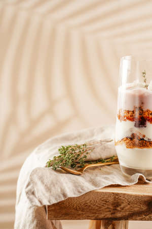 Tall glass with a layered creamy yoghurt dessert trifle with cookies, amaretti, cherries and thyme on a linen cloth on a rustic wooden table at a tropical settingの写真素材