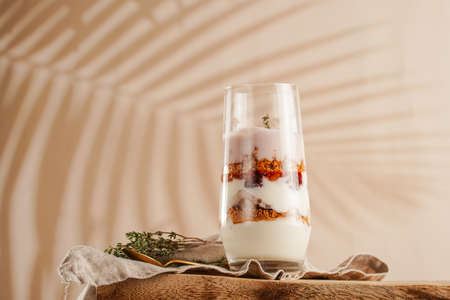 Tall glass with a layered creamy yoghurt dessert trifle with cookies, amaretti, cherries and thyme on a linen cloth on a rustic wooden table at a tropical settingの写真素材