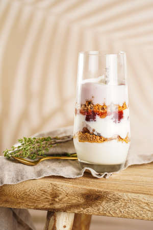 Tall glass with a layered creamy yoghurt dessert trifle with cookies, amaretti, cherries and thyme on a linen cloth on a rustic wooden table at a tropical settingの写真素材