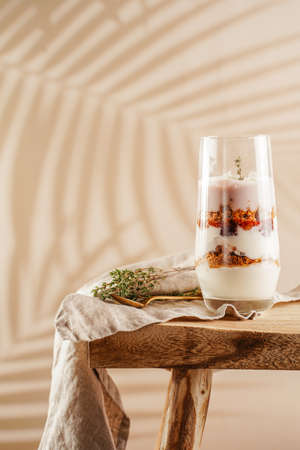 Tall glass with a layered creamy yoghurt dessert trifle with cookies, amaretti, cherries and thyme on a linen cloth on a rustic wooden table at a tropical settingの写真素材