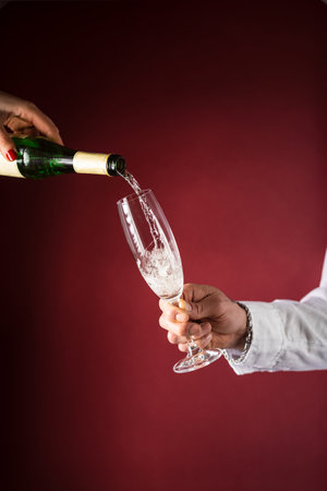 Male hand holding a champagne glass with champagne and female hand pouring champagne from bottle with many drops dripping on burgundy backgroundの写真素材