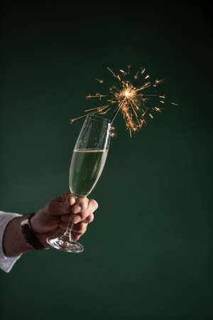 A male hand in white shirt holding glass of sparkling wine champagne with sparkler in it on dark green backgroundの写真素材