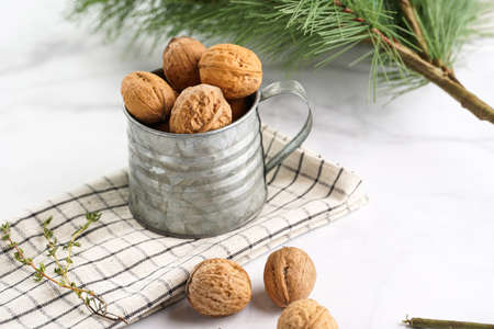 A metal mug with whole brown walnuts on a checkered linen cloth and a christmas fair-tree branch on...
