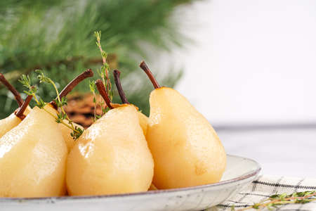 Peeled poached pears on a white oval plate on a checkered linen cloth and a christmas fair-tree branch on a white table surfaceの写真素材