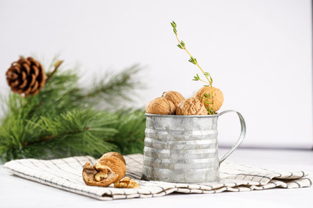 A metal mug with whole brown walnuts on a checkered linen cloth and a christmas fair-tree branch on a white table surfaceの写真素材
