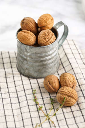 A metal mug with whole brown walnuts on a checkered linen cloth on a white table surfaceの写真素材